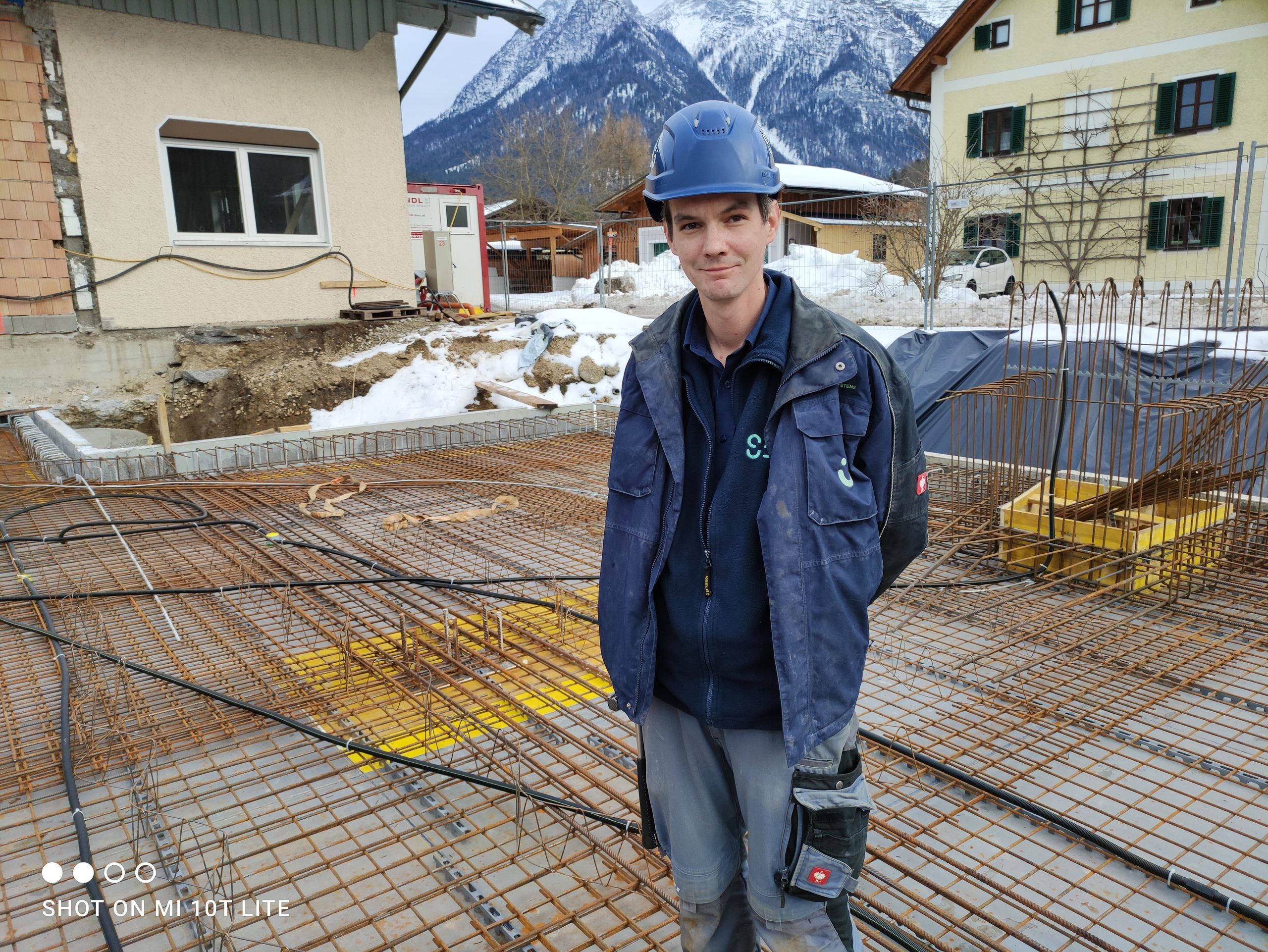 SES-Monteur mit blauem Bauhelm steht auf der Rohbaudecke einer Baustelle im Salzkammergut, im Hintergrund verschneite Berge.