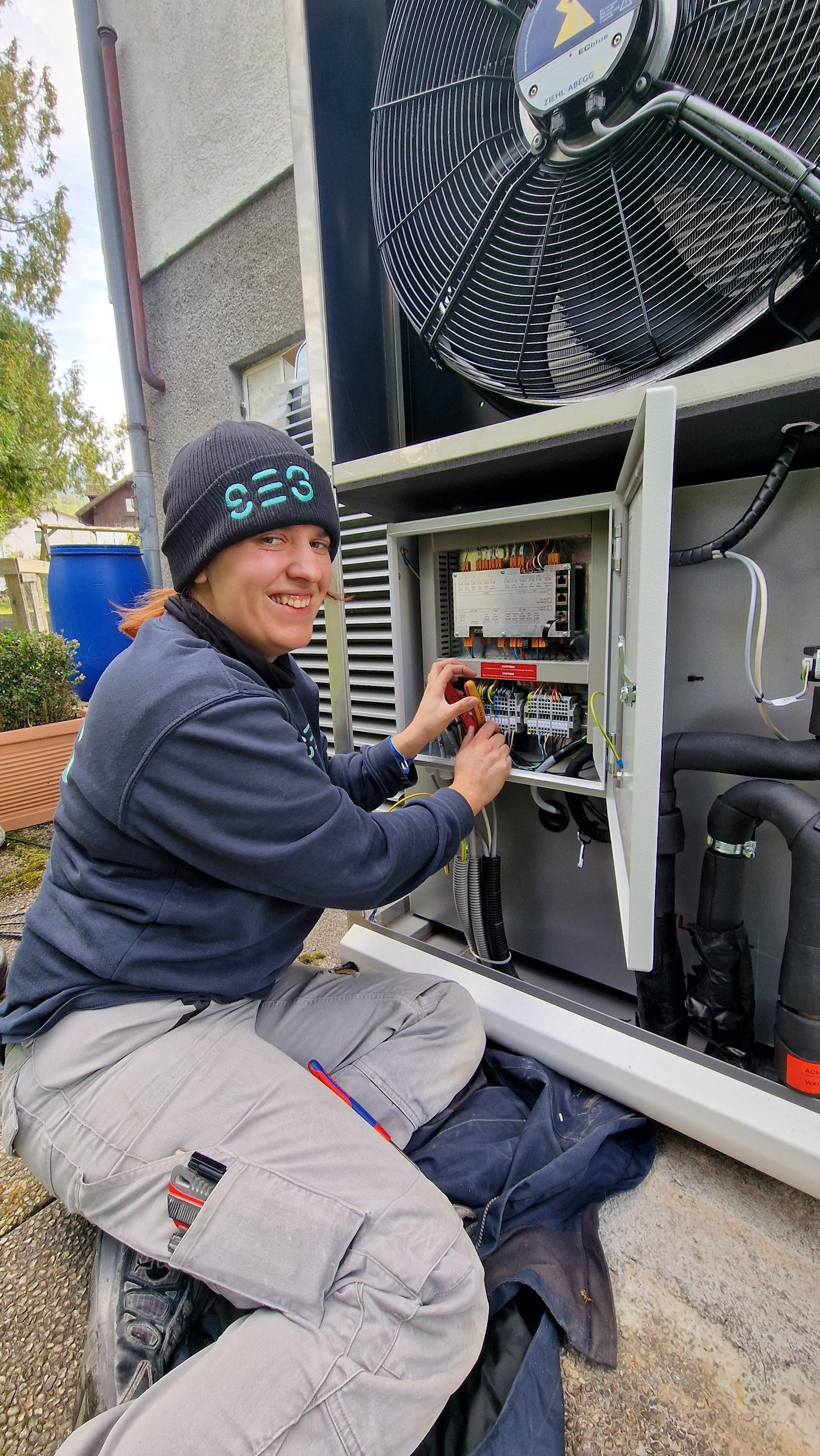 SES-Monteurin mit SES-Beanie arbeitet am geöffneten Schaltschrank einer Wärmepumpe im Freien.
