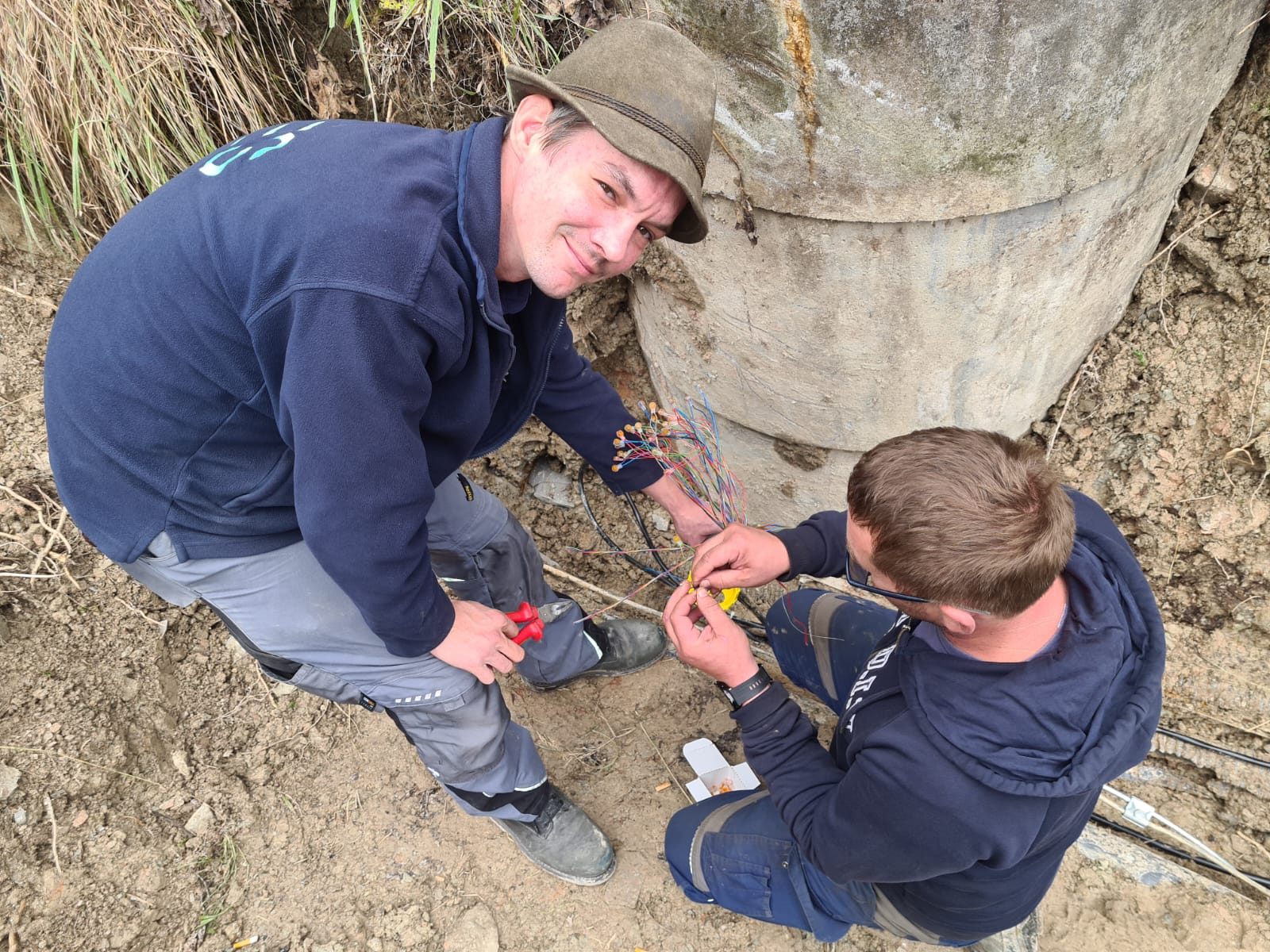 Zwei SES-Elektriker verbinden im Kabelgraben an einem Betonfundament die Adern eines Erdkabels.